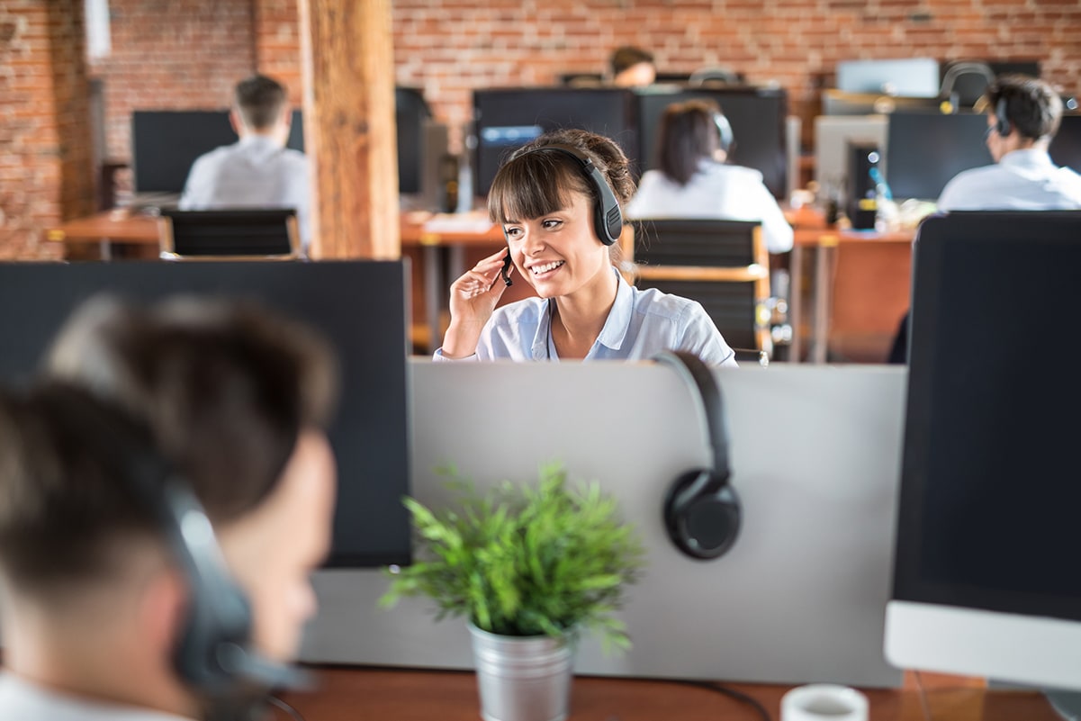 An image of the inside of a call center. Focus is on a women with headphone smiling into the microphone.