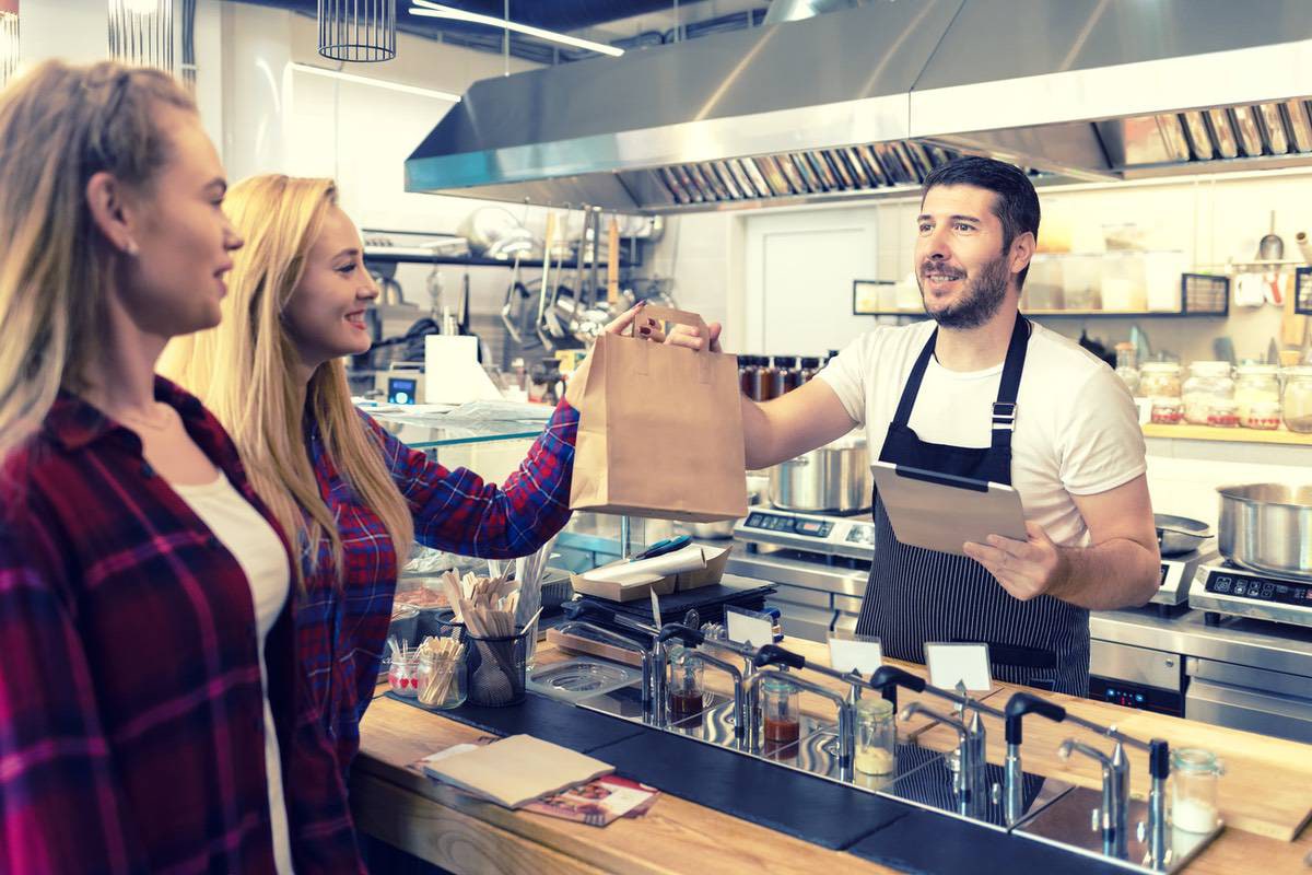 A man behind a food counter hands a brown paper bag to a woman